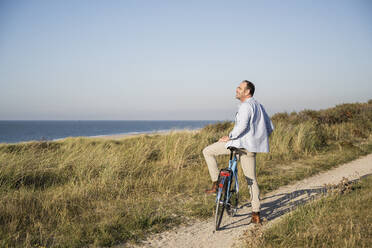 Smiling man looking at view while standing with bicycle on beach against clear sky - UUF21979