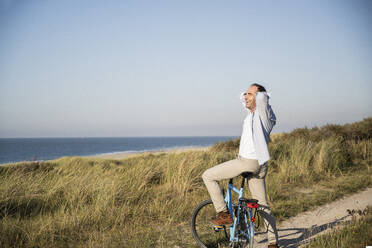 Carefree man on bicycle with hands behind head at beach against clear sky - UUF21980