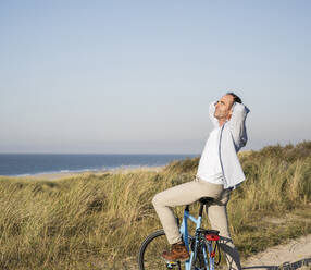 Mature man on bicycle with hands behind head at beach against clear sky - UUF21981