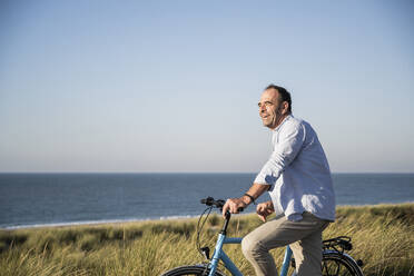 Happy mature man with bicycle at beach against clear sky - UUF21982