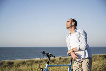 Mann mit geschlossenen Augen lehnt sich am Strand gegen den klaren Himmel auf ein Fahrrad - UUF21984