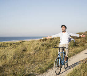 Unbekümmerter Mann mit ausgestreckten Armen auf dem Fahrrad am Strand gegen den klaren Himmel - UUF21987