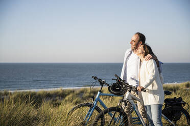 Happy mature couple with bicycles at beach against clear sky during weekend - UUF21988