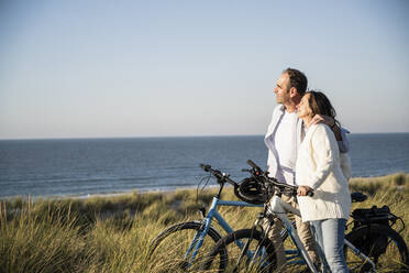 Smiling couple with bicycles looking at view while standing on beach against clear sky during weekend - UUF21989