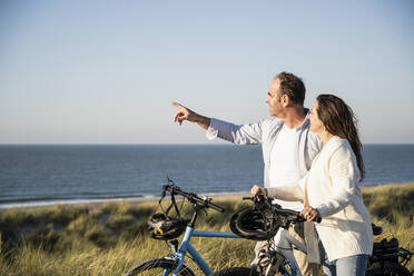 Happy couple with bicycles looking at view while standing on beach against clear sky - UUF21990