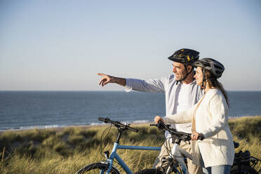 Smiling couple in cycling helmets looking at view while standing on beach against clear sky - UUF21991