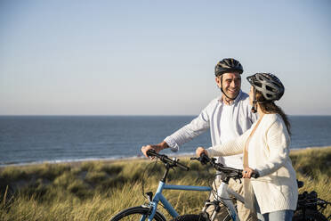 Happy couple in cycling helmets standing with bicycle at beach against clear sky - UUF21992