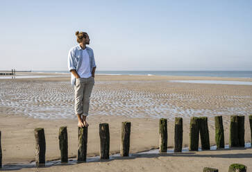 Junger Mann, der auf einem Holzpfosten am Strand steht und die Aussicht gegen den klaren Himmel betrachtet - UUF21997