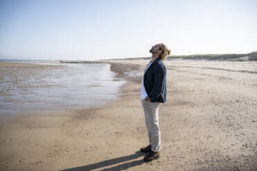 Junger Mann steht mit den Händen in den Taschen am Strand gegen den klaren Himmel - UUF21999