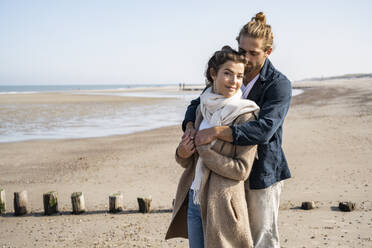 Junges Paar, das sich am Strand umarmt, gegen den klaren Himmel stehend - UUF22003