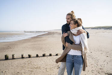 Young couple looking away while embracing at beach against clear sky - UUF22005