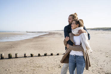 Junges Paar mit geschlossenen Augen umarmt, während am Strand gegen den klaren Himmel stehen - UUF22006