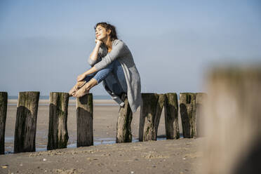 Nachdenkliche junge Frau sitzt auf Holzpfosten am Strand gegen den Himmel - UUF22008