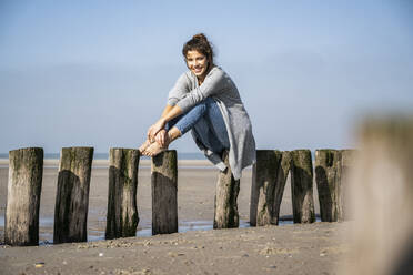 Smiling young woman sitting on wooden posts at beach against sky during sunny day - UUF22009