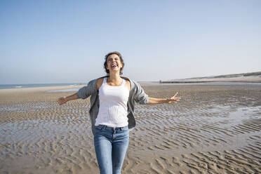 Fröhliche junge Frau mit ausgestreckten Armen, die am Strand gegen den klaren Himmel spazieren geht - UUF22012