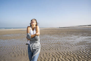 Happy young woman hugging self while standing at beach against clear sky - UUF22017