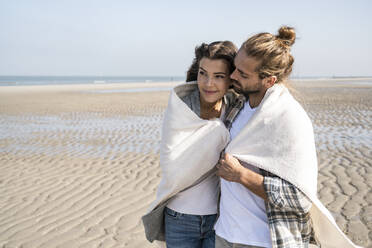 Romantisches junges Paar in Decke gehüllt am Strand stehend - UUF22021