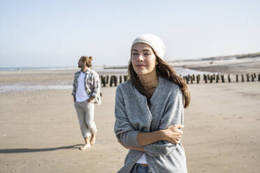Junge Frau im Stehen mit geschlossenen Augen am Strand, Mann im Hintergrund - UUF22024