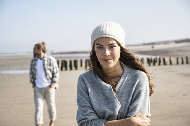 Lächelnde junge Frau im Stehen mit verschränkten Armen am Strand, während Mann im Hintergrund - UUF22025