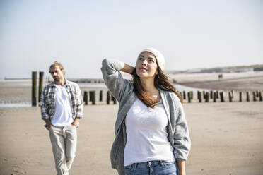 Nachdenkliche junge Frau schaut auf, während ein Mann im Hintergrund am Strand spazieren geht - UUF22026