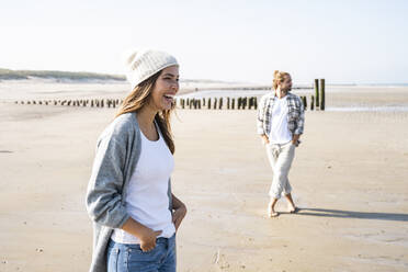 Fröhliche junge Frau mit den Händen in den Taschen am Strand stehend an einem sonnigen Tag - UUF22029