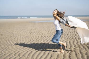 Carefree young woman running while holding blanket at beach during sunny day - UUF22031