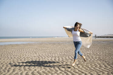 Unbeschwerte junge Frau, die am Strand gegen den klaren Himmel läuft und eine Decke hält - UUF22032