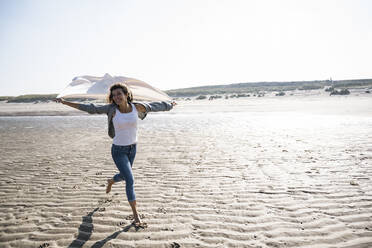 Unbekümmerte Frau, die an einem sonnigen Tag am Strand läuft und eine Decke hält - UUF22033