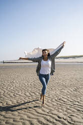 Cheerful woman running while holding blanket at beach during weekend - UUF22034