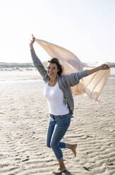 Unbeschwerte junge Frau, die an einem sonnigen Tag am Strand läuft und eine Decke hält - UUF22035