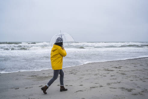 Woman in yellow raincoat walking with umbrella at sea shore against sky - UUF22038