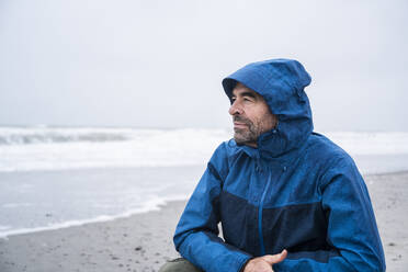 Nachdenklicher reifer Mann in blauem Regenmantel, der am Strand vor dem Himmel kauert - UUF22041