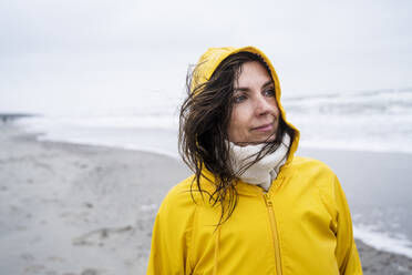 Thoughtful mature woman in yellow raincoat standing at beach against sky - UUF22043