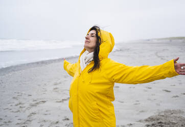 Mature woman in yellow raincoat standing with arms outstretched at beach - UUF22045