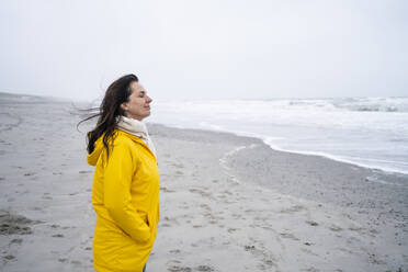 Lächelnde reife Frau steht mit Händen in den Taschen am Strand gegen den Himmel - UUF22046