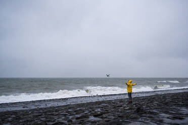 Woman with arms outstretched standing at sea shore against cloudy sky - UUF22048