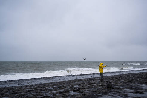 Woman with arms outstretched standing at sea shore against cloudy sky - UUF22048