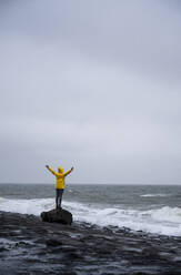 Woman with arms outstretched standing on rock at beach against cloudy sky - UUF22050