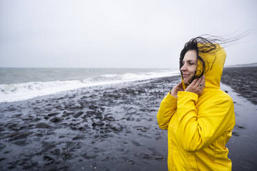 Smiling mature woman in yellow raincoat looking at view while standing on sea shore - UUF22052