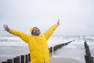 Unbekümmerter reifer Mann im Regenmantel, der schreiend mit erhobenen Armen am Strand vor dem Himmel steht - UUF22056