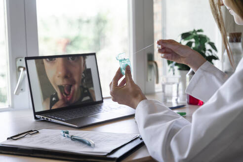 Young female dentist showing dental floss to patient through video call on laptop at home - AFVF07421