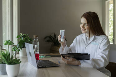 Smiling female dentist showing toothpaste to patient through video call on laptop at home - AFVF07426