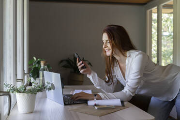 Smiling psychologist using mobile phone while working on laptop at home - AFVF07429