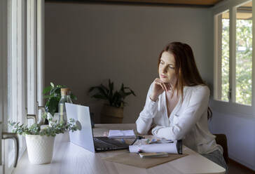 Thoughtful psychologist looking at laptop while sitting with hand on chin at home - AFVF07430