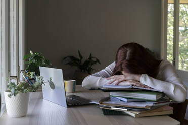 Tired doctor leaning on stack of book while sitting at home - AFVF07439