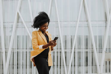 Smiling female entrepreneur using smart phone listening to music through earphones standing against building - GMLF00756