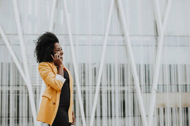 Smiling female entrepreneur talking on smart phone while standing against building - GMLF00758