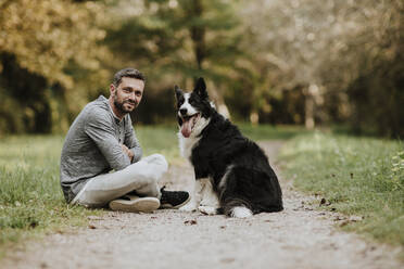 Smiling man and dog sitting on footpath at public park - GMLF00782