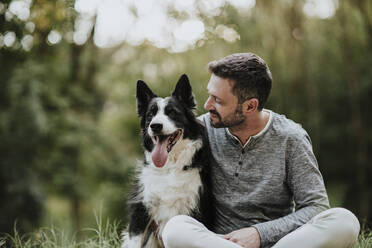 Lächelnder Mann, der seinen Hund anstarrt, während er in einem öffentlichen Park sitzt - GMLF00786