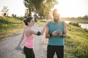 Männlicher Sportler, der sein Smartphone benutzt, während eine Sportlerin mit hochgelegtem Bein im Park bei Sonnenuntergang steht - SBAF00052
